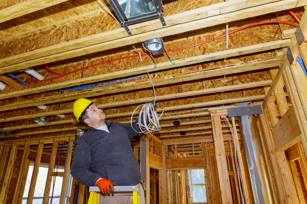 Electrician installing electrical wiring in new home construction, running cables through studs and connecting junction boxes while wearing safety gear